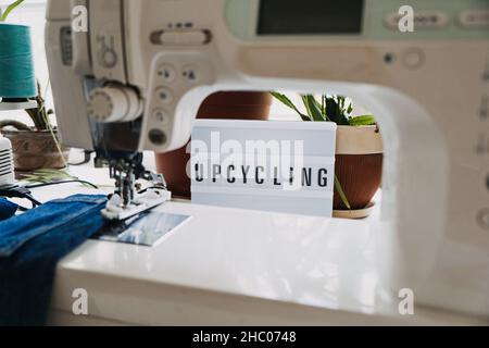Upcycling text on light board on sewing machines with old blue jeans background. Denim clothes, scissors, thread and sewing tools in sewing studio Stock Photo
