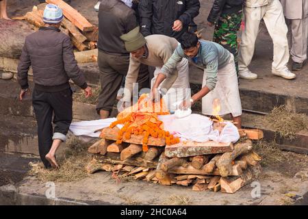 Workers remove the offerings from the body as the body starts to burn in the cremation ceremony.  Pashupatinath, Kathmandu, Nepal. Stock Photo