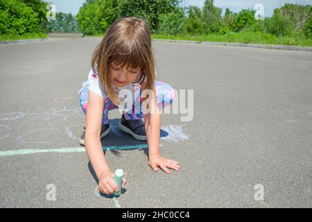 A little girl draws with chalk on an asphalt road. Concept - children's drawings of a picture on asphalt. Stock Photo