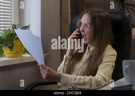 Angry woman boss talking on  phone and shouting, holding documents. Director scolds subordinates on smartphone Stock Photo