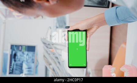 Vertical video: Woman holding smartphone with horizontal green screen at oral care clinic. Dentist looking at chroma key with isolated background and mockup template for dental checkup Stock Photo
