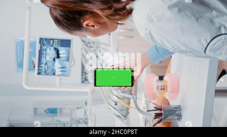 Vertical video: Woman vertically holding mobile phone with green screen in dental cabinet. Stomatologist looking at chroma key and isolated mockup template on smartphone for oral care at office Stock Photo