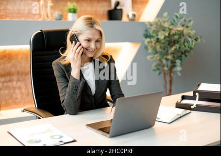 Busy friendly successful intelligent Caucasian businesswoman, freelancer or top manager sitting at table in the office with laptop, talking to client or employees on mobile phone, smiling Stock Photo