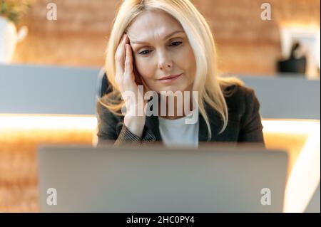 Sleepy emaciated senior woman, business woman or manager, in stylish suit, tired of working at laptops, experiencing stress and headache, massaging her temple, needs rest, looking languidly at screen Stock Photo