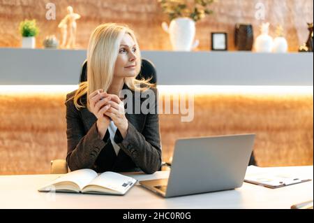 Concentrated serious senior woman, ceo, manager or broker sitting at table in modern office wearing formal suit, pondering business idea or financial strategy, is looking to the side thoughtfully Stock Photo