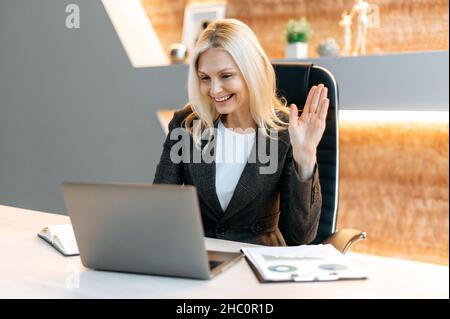 Positive beautiful influential Caucasian mature woman, top company manager, sits in office using laptop, talking online with client or employees, discussing project, holds consultation, waving hand Stock Photo