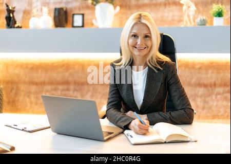 Confident intelligent caucasian middle aged business woman, financial manager, broker, in formal suit, sitting at table in office, using laptop, taking notes, looking at camera, smiling friendly Stock Photo