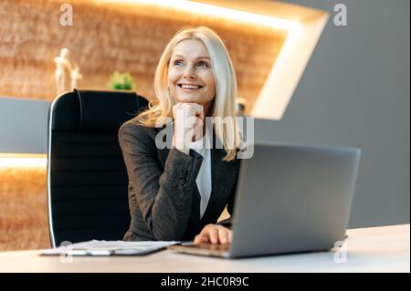 Happy joyful successful blonde caucasian mature business woman, manager, finansist, sitting at table in formal clothes, taking break from work, looking pensively to side, dreaming about vacation Stock Photo
