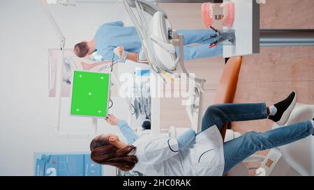 Vertical video: Dentist and nurse analyzing teeth x ray and talking about dental checkup, working with horizontal green screen on monitor. Specialist and man looking at radiography and chroma key display Stock Photo