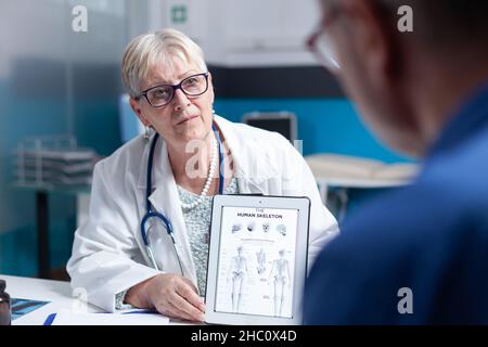 Close up of medic holding tablet with image of human skeleton to explain diagnosis to old man. Doctor and patient looking at bones and spinal cord illustration on device for examination. Stock Photo