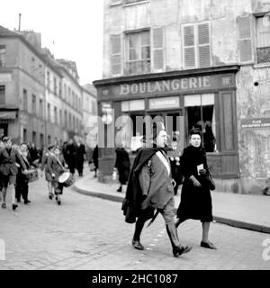 A procession in 1945 in Paris Montmartre comes from the direction of Sacre-Cœur walking down Rue Norvins and turning left down Place Jean-Baptiste Clement. Leading is an elderly man wearing a Napoleon hat, a cloak, and riding boots. Cigar in mouth, a sword dangles to his left side. He appears to have lost his right arm. Behind him in the march are a group of boys beating drums. A US Navy enlisted man watches from one side and A woman watches and then joins at the front of the group. The Consulat d’Auvergne and Restaurant L’ambassade and Cafes Gilbert are in view Stock Photo