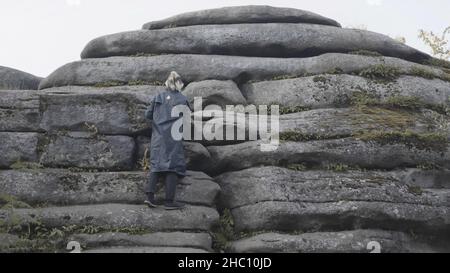 Rear view of a man in a long jacket and colored hair trying to climb a stony mountain. Male hiker climbing giant bouldrers on cloudy sky background. Stock Photo