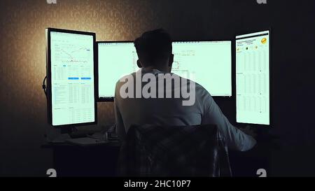 Back view of modern programmer sitting and writing code in dark room. Young man playing computer games in dark room. Stock Photo