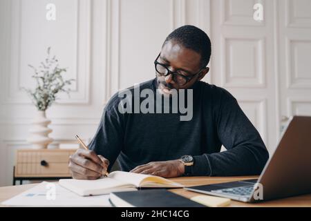 Stunning concentrated Aframerican male lawyer in casual wear making notes in agenda Stock Photo