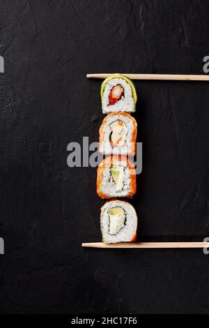 Four sushi rolls with salmon, eel, shrimp, avocado on black surface between chopsticks Stock Photo