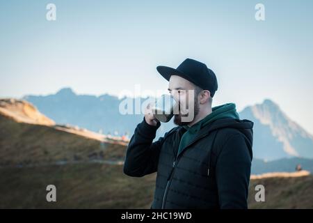 Bearded traveler man drinks tea or coffee from  metal cup while resting on a rock on a beautiful lanscape background Stock Photo