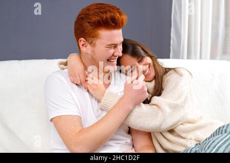 Cheerful married couple resting on couch enjoy lazy weekend together at home having fun over white and grey background. Stock Photo