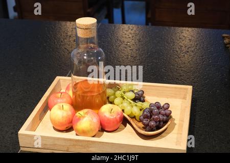 Flacon of homemade vinegar on a wood serving tray with apples and grapes, on a modern black stone kitchen counter Stock Photo