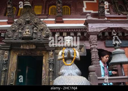 A woman about to ring a bell next to the brass statue of a former king, in Hiranya Varna Mahavihar (Kwa Bahal, Golden Temple), Patan (Lalitpur), Nepal Stock Photo