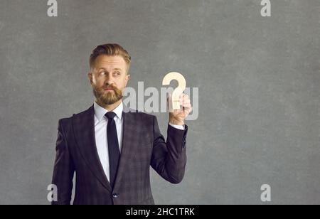 Confused puzzled businessman holding wooden question mark standing on gray on background. Stock Photo