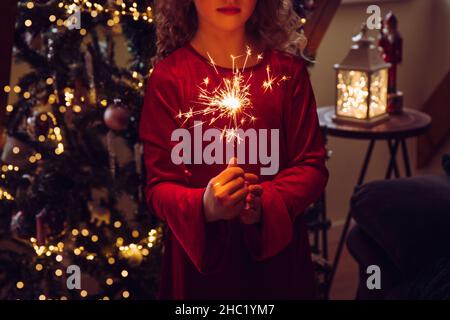 Cute girl child holding sparkler candle in hands in home living room, decorated Christmas tree on background, dark room, lit by sparkler candle. Happy Stock Photo