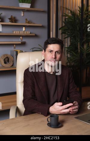 Young serious man in a burgundy suit with a mobile phone sit in an office at a table and looks at the camera. Dark hair, brown eyes, white skin. In th Stock Photo