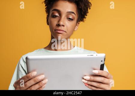 Young black woman looking at tablet computer while holding gadget in hands isolated over yellow background Stock Photo