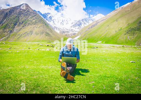 Middle age male person with white full cap sits with crossed legs on the grass with laptop on his knees and types with smile in his face. Remote work Stock Photo