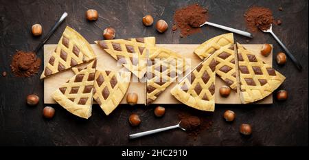 Crostata, eight slices of hazelnut and cocoa tart on wooden cutting board with spoons and cocoa powder. Top view on brown plaster background, flat lay Stock Photo