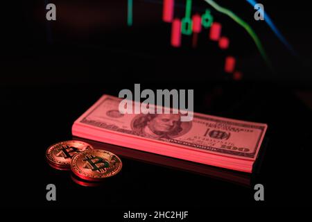 2 bitcoin coins are lying next to stack of banknotes and in the background is a falling bitcoin chart Stock Photo