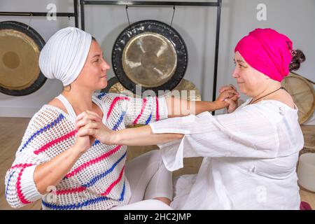 Two women dressed in white with turban doing kundalini yoga sitting on the floor holding their hands Stock Photo