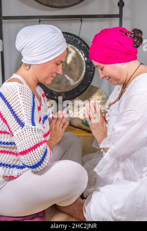 Women dressed in white with turban practicing kundalini yoga sitting on the ground one in front of the other and hands together Stock Photo