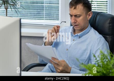 Focused office worker reads documents and gnaws on arch of glasses. Male accountant. Assistant Director Stock Photo
