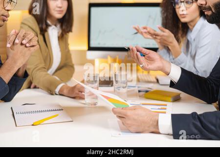 Glad to hear it. Portrait of optimistic young qualified manager is looking at his colleagues with smile while sitting at table and having pleasant communication. Man holding diagrams Stock Photo