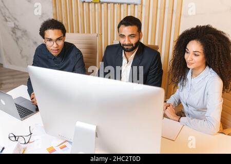 Diverse work team having virtual meeting with outsource team. Multiracial colleagues sitting at the desk in modern office space and talking via video call with group on the computer screen Stock Photo