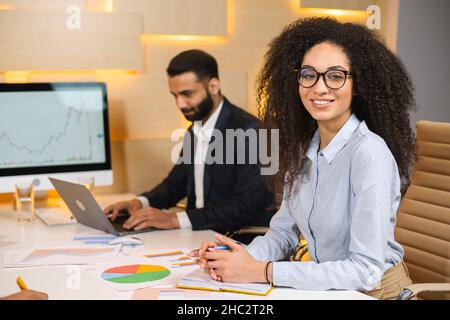 Nice to work with you. Portrait of cheerful young female manager in glasses is sitting at the table and posing to the camera. Her male colleague is sitting at the background Stock Photo