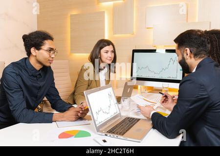 Millennial colleagues sitting at office desk, looking at computer and talking about business issues. Diverse employees communicate near each other and negotiating on projects together Stock Photo