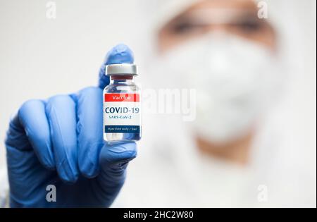 Medical NHS worker holding COVID-19 SARS-CoV-2 vaccine glass ampoule vial,wearing personal protective gear,white clean suit,blue gloves,Coronavirus cu Stock Photo