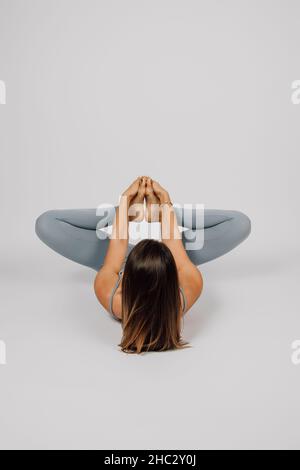 Woman does sports. Brunette in gray sportswear does exercise from yoga or fitness, she lies on her back in frog pose with her legs bent at knees, rear Stock Photo