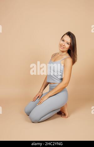 Brunette poses on neutral background. Slender young woman in gray top and leggings smiles and stands, leaning on her knees and putting her hands on Stock Photo