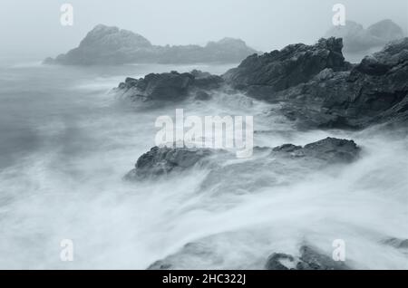 A serene coastal scene shrouded in mist featuring a series of rugged rocks partially submerged in crashing waves at Big Sur, California. Stock Photo