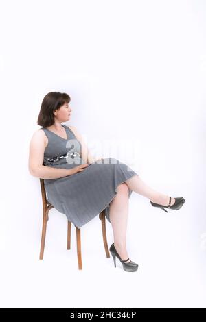 Plus size women in gray dress and heels sits on chair. Isolated on a white background Stock Photo