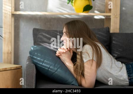 Teenage girl lies on couch and looks dreamily in front of her. Young woman is resting on sofa in living room. Front view. Stock Photo