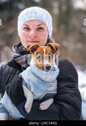 Young woman in winter jacket holding her Jack Russell terrier dog wearing warm winter clothing on hands, blurred snow covered trees background Stock Photo