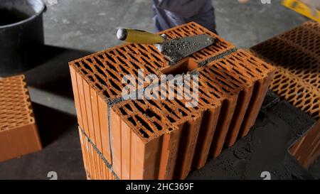 Bricklayer worker putting away extra cement with trowel putty knife on grey floor background. Man in gloves building a red brick wall. Stock Photo