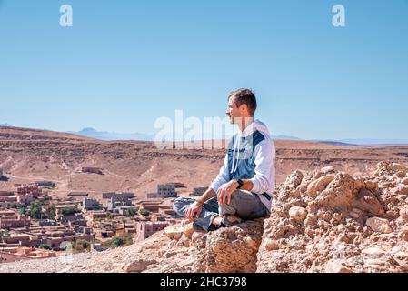 Young man admiring view while sitting on rock against deserted land and sky Stock Photo