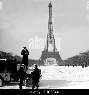 US Navy men and jeep in snow in the Champ de Mars with the Paris Eiffel Tower, 1945. In perhaps the first heavy snow after Liberation, three American navy men have pulled their jeep into the Champs de Mars with the Eiffel Tower in the background. No other people are in sight. The tower is depicted from base to top and the Palais de Chaillot can be seen between the legs. A US Chief Petty Officer in peacoat stands on the hood of the jeep with a movie camera. Two seamen in navy caps and peacoats hold a map of Paris. Stock Photo