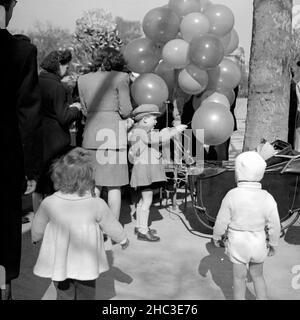 Two photographs of a scene where children are getting balloons in an unidentified Paris park in 1945. One photograph shows three parents, three children standing, and a partially obscured baby in a stroller. The other photo shows a smiling balloon-man (slightly out of focus) surrounded by several men. Some of the men wear uniforms. Stock Photo