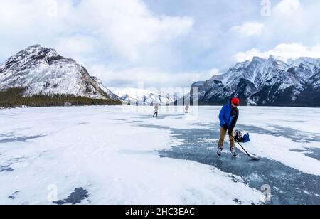 Boy practicing ice hockey on frozen Lake Minnewanka in the Canadian Rockies of Banff National Park during the cold Winter with unidentifiable visitors Stock Photo