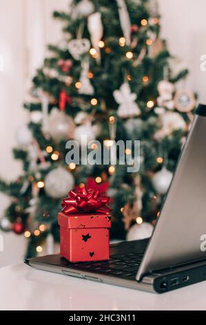 Small red present with gift bow next to laptop on table in front of decorated christmas tree Stock Photo